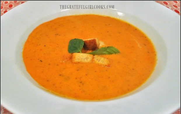 Creamy tomato basil soup is served in white bowl, with croutons and basil leaves as garnish.