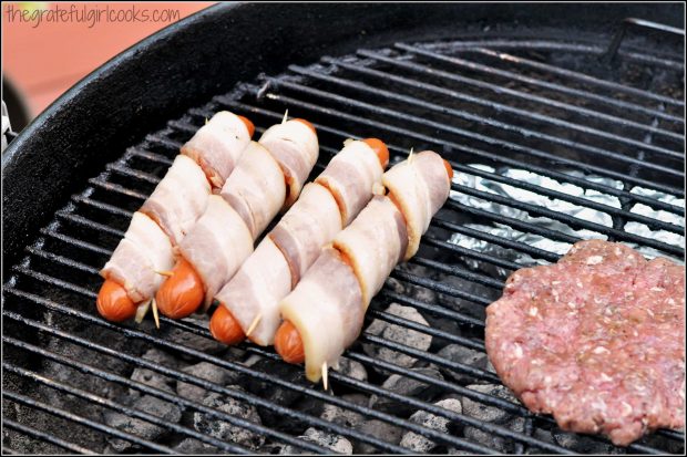 Four hot dogs wrapped in bacon cooking over hot coals.