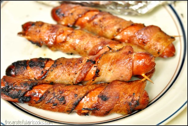 Four bacon-wrapped grilled hot dogs on plate, ready to eat.