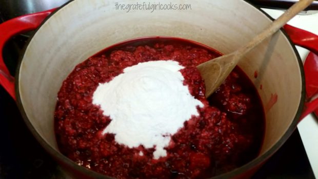 Fruit pectin powder is added to crushed raspberries in large pot.