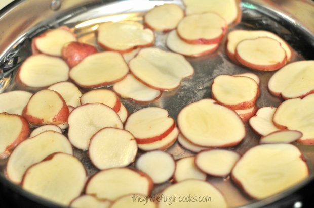 Red potato slices begin cooking in a bit of water, in a large skillet.