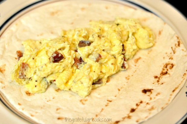 Scrambled eggs with bacon is placed in center of warm flour tortilla for breakfast burritos.