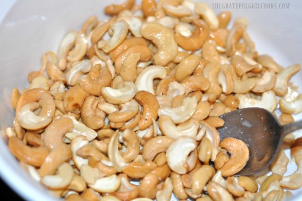 Melted coconut oil is stirred into cashews in bowl.