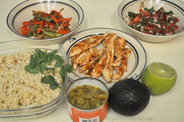 Ingredients (peppers, salsa, rice, chicken, avocado, chiles, and lime) are ready to build the chicken fajita rice bowl.