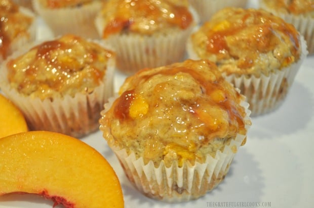 Close up photo of one peach jam muffin, with fresh peach slice in front.