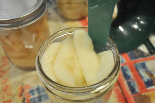 Air bubbles are removed from can of pears by inserting plastic utensil into jar.