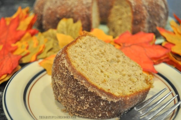 Apple Cider Bundt Cake slice on plate with fork, with rest of cake in background with Fall leaves.