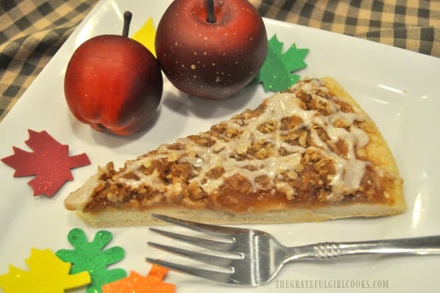 A slice of apple pie dessert pizza on white plate, with apple and leaf decorations.