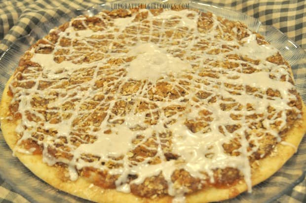 Apple pie dessert pizza is decorated with cinnamon glaze before slicing.