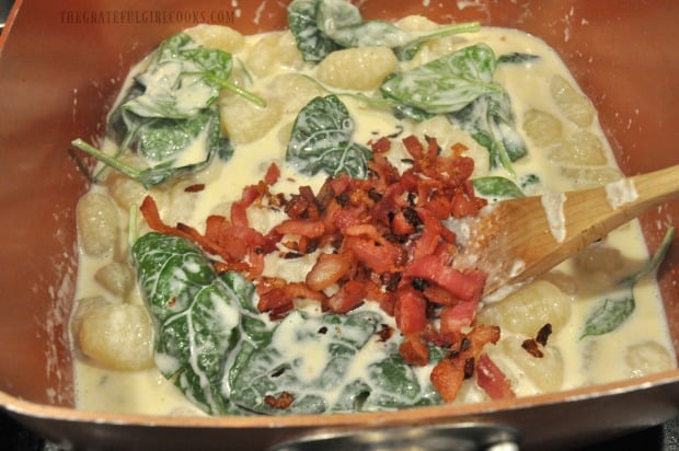Cooked gnocchi, sauce, fresh spinach and bacon is mixed for bacon parmesan spinach gnocchi.