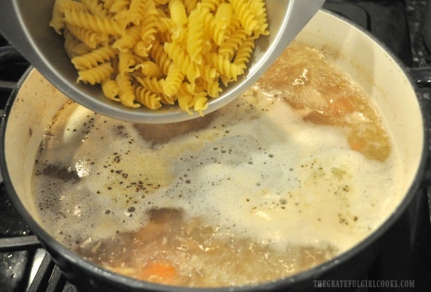 Uncooked rotini pasta is added to boiling chicken noodle soup in pan. 