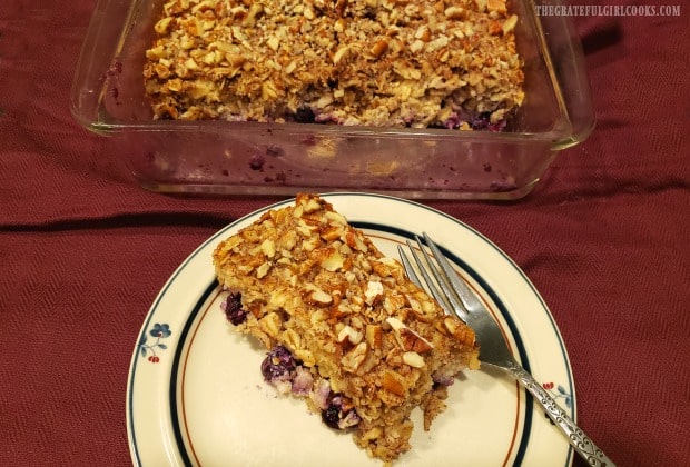 A slice of blueberry baked oatmeal on a plate, with rest of dish in background.
