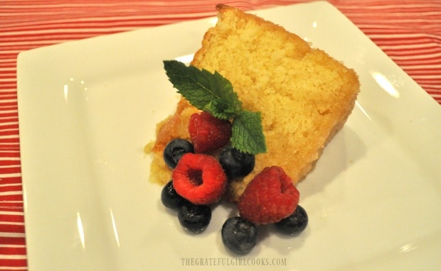 A slice of glazed butter cake on a plate with fresh blueberries, raspberry and a mint sprig.