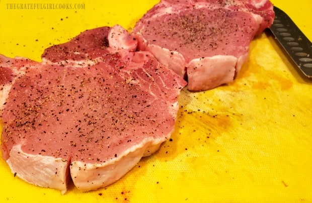 Pork chops are seasoned with salt and pepper, and fat is sliced through on outside of each one.