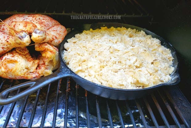 A cast iron skillet full of cobbler is placed onto a Traeger grill, next to roasting chicken.