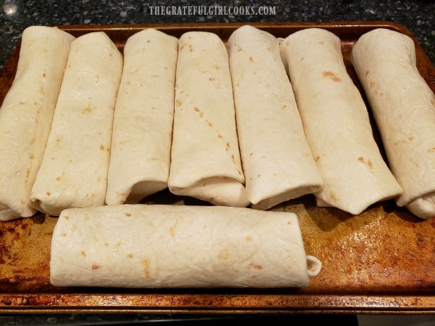 Eight rolled chicken chimichangas on pan are ready to cook.