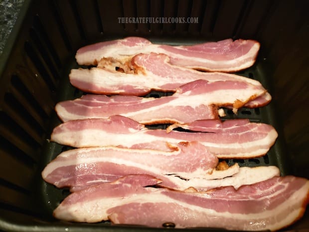 The bacon strips are laid side by side in the basket of the air fryer to cook.