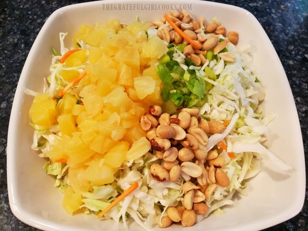 Pineapple tidbits, green onions, peanuts are added to the coleslaw mix in bowl.
