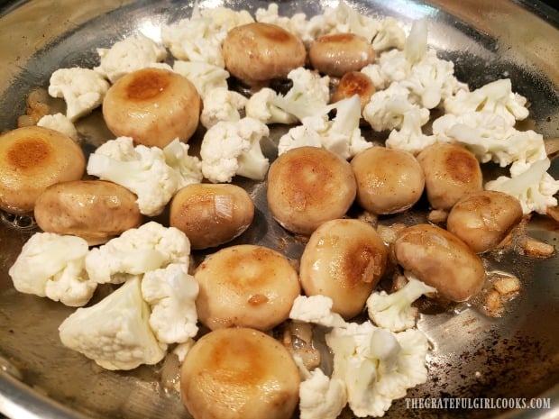 Cauliflower florets are added to the mushrooms in the skillet.
