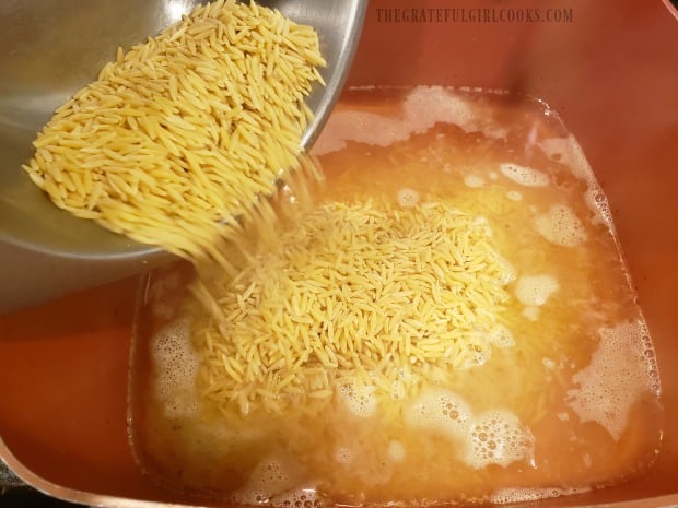 Uncooked orzo pasta is added to boiling chicken broth in pan, to cook.