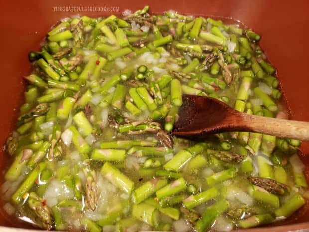 Asparagus, chicken broth, and red pepper flakes are added to the cream of asparagus soup in pan.