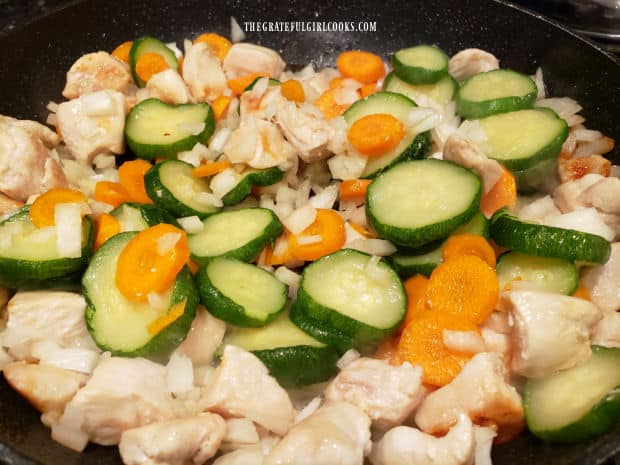 Garlic, onion, zucchini and carrot slices are added to the cooking chicken pieces.