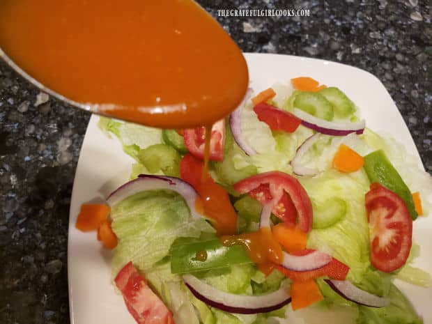 A Tablespoon of salad dressing is drizzled onto a mixed green salad on a white plate.