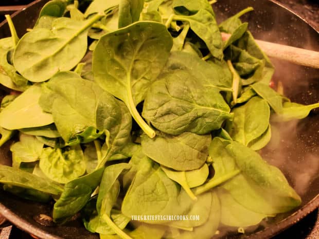 Baby spinach leaves are added to the cooked bacon in a large skillet.