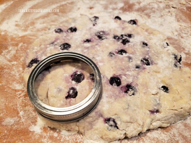 A small canning jar lid is used to cut out 8 blueberry biscuits from the dough.