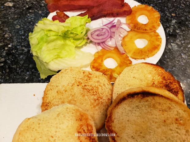 Toasted hamburger buns, bacon, lettuce, onion and grilled pineapple ready for burgers.