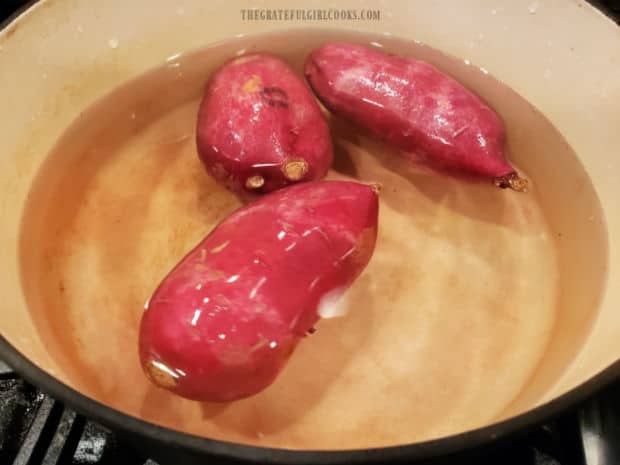 Three medium sized sweet potatoes cooking in boiling water in a large saucepan.