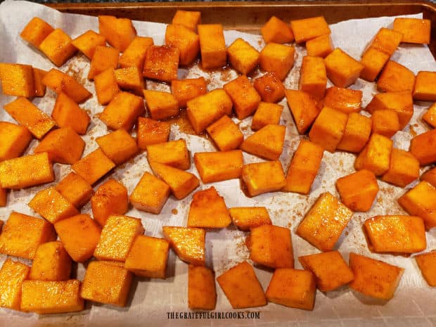 Seasoned maple butternut squash cubes are spread in single layer on parchment paper for baking.