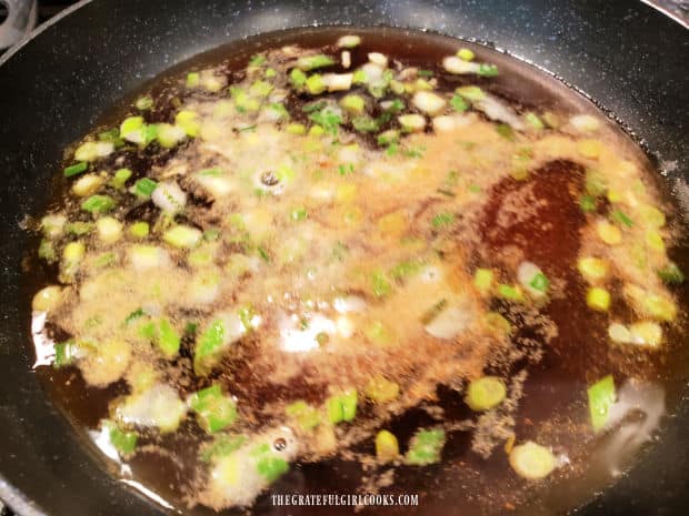 Water, soy sauce, and spices are added to the cooked green onions in the skillet.