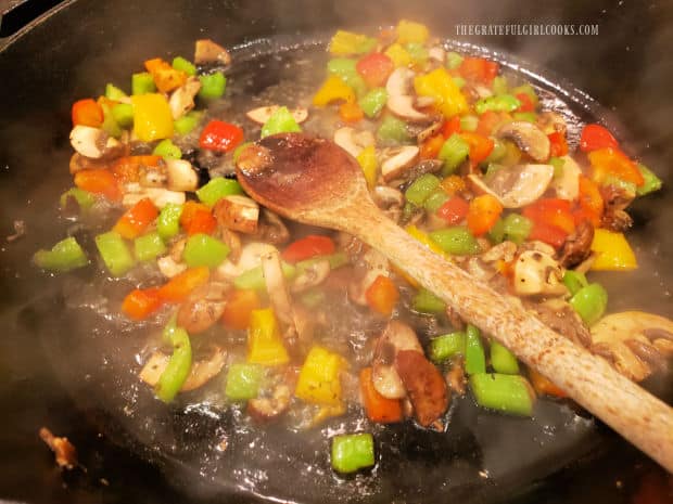 Water is added to the stir-fried veggies in skillet with a wooden spoon.