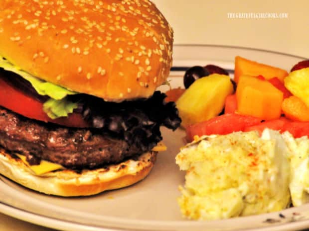 A hamburger made with one of the Cajun-seasoned burger patties, on plate with fruit and potato salad.