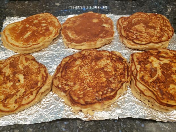 Six chunky apple cinnamon pancakes cool on aluminum foil after being cooked.
