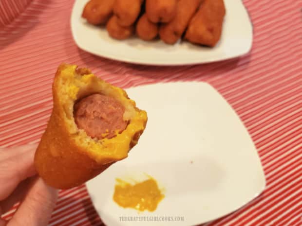 Corn dog, dipped in mustard, with a plate of corn dogs in background.