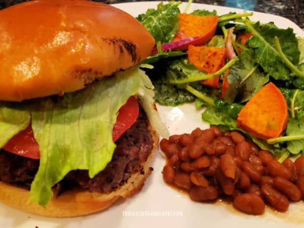 A white plate with a hamburger, baked beans and kale and sweet potato salad.