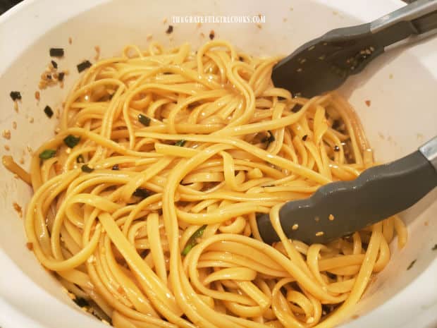 Tongs being used to combine Asian sauce and hot linguini pasta in a bowl.
