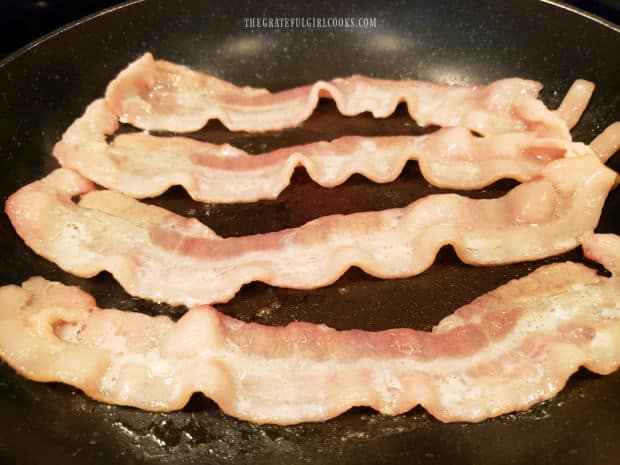 Several strips of bacon cooking in a large black skillet.