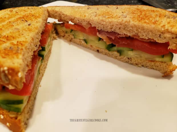 A Bacon Tomato Cucumber Sandwich is sliced in half, and served on a white plate.