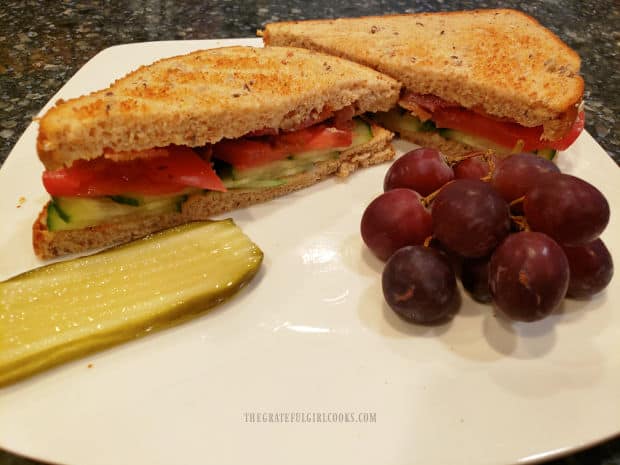 A pickle slice and red grapes are served on a plate with the Bacon Tomato Cucumber Sandwich.