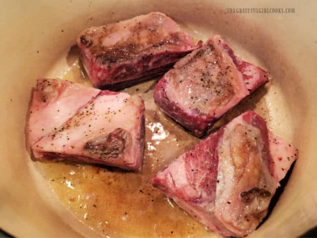 Four beef short ribs are browned in hot oil in a large Dutch oven.
