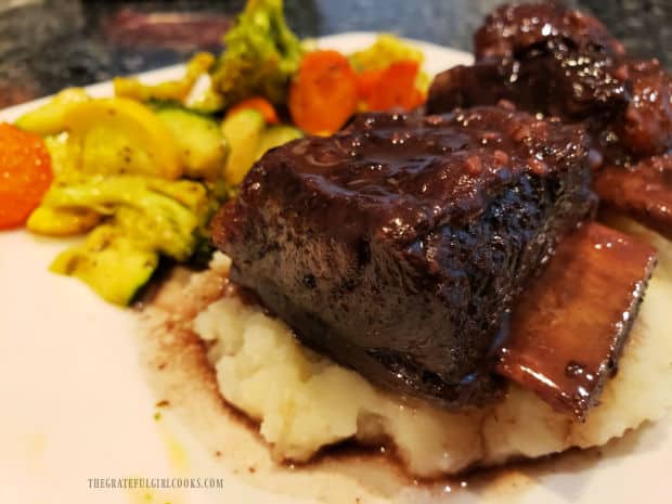 One of the beef short ribs on mashed potatoes, with veggies in the background.