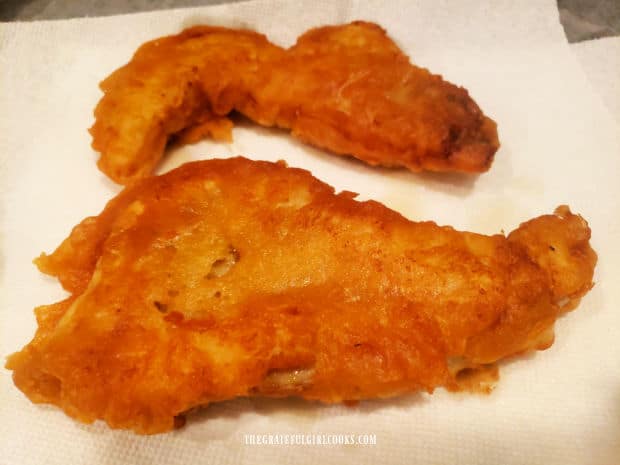 Two batter-fried rockfish fillets drain on paper towels after cooking.