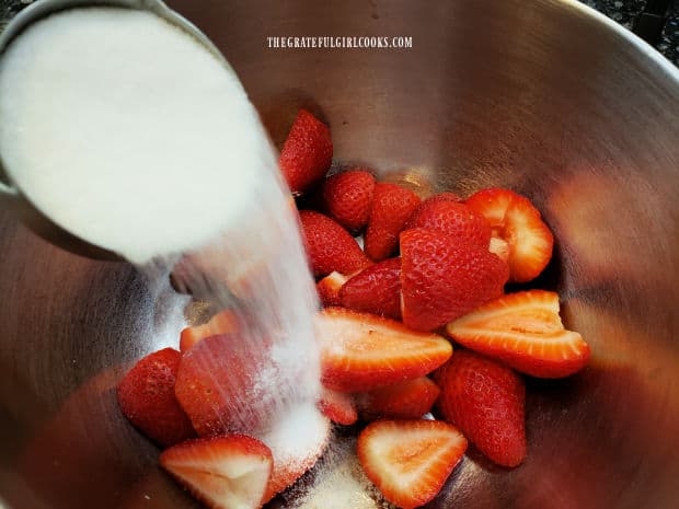 Granulated sugar is added to the halved strawberries in a bowl.