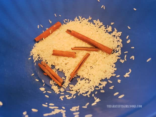 Two cinnamon sticks (broken in pieces) in a blue bowl with uncooked rice.