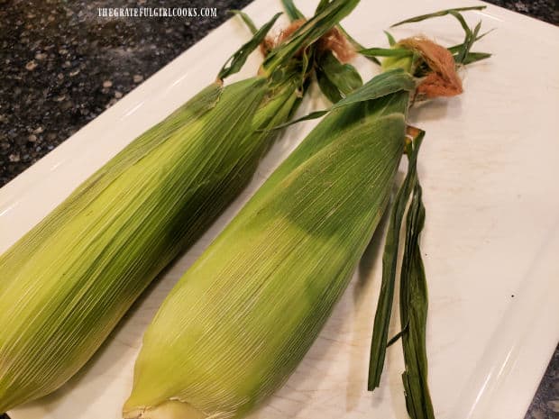 Two ears of fresh corn, with the outer husks intact.