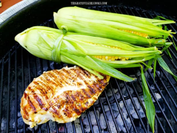Grilled unshucked corn on the cob, cooking on BBQ by a piece of chicken.
