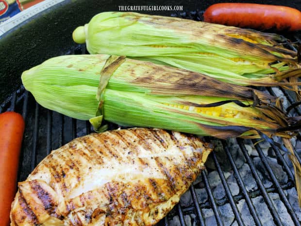 Two pieces grilled unshucked corn on the cob, with chicken and hot dogs on BBQ.